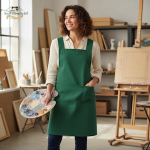 Può includere: Un grembiule da artista verde scuro con un'ampia tasca frontale e scollatura quadrata. Il grembiule è indossato sopra una camicia bianca abbottonata. L'artista tiene in mano una tavolozza di legno con vari colori di vernice. Il logo "SewPatternByAdil" è nell'angolo in alto a sinistra.