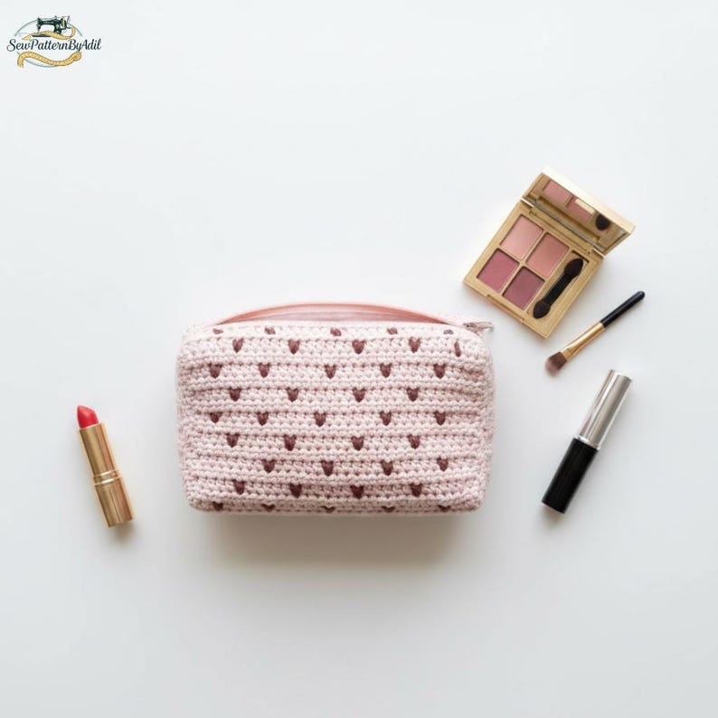 May include: A light pink crochet cosmetic bag with a heart pattern, a gold lipstick, a makeup palette, a brush, and a mascara tube on a white surface. The bag has a pink zipper.