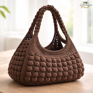 Può includere: Una borsa a mano all'uncinetto marrone fatta a mano con un motivo a punto nocciola strutturato. La borsa ha una forma arrotondata con un manico intrecciato. La borsa è su una superficie di legno. Il testo "SewPatternByAdil" è nell'angolo in alto a destra.