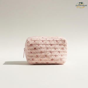 May include: A light pink, crocheted pouch with a zipper closure. The pouch is decorated with small, evenly spaced pink hearts. The item is handmade and appears to be a cosmetic bag or small storage container.