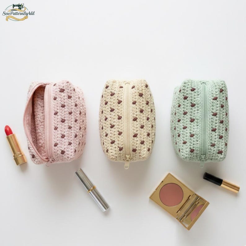 May include: Three handmade crochet cosmetic bags in pink, cream, and mint green, each with a brown dot pattern and a zipper closure. Makeup items, including lipstick and blush, are arranged around the bags. The bags are approximately 6 inches tall.