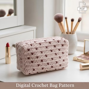 Può includere: Una borsa all'uncinetto rosa chiaro con chiusura a cerniera, ornata da piccoli motivi a forma di cuore marrone scuro. La borsa è esposta su una superficie bianca, insieme a pennelli per il trucco, rossetto e un specchietto compatto. Il testo in basso recita "Digital Crochet Bag Pattern."