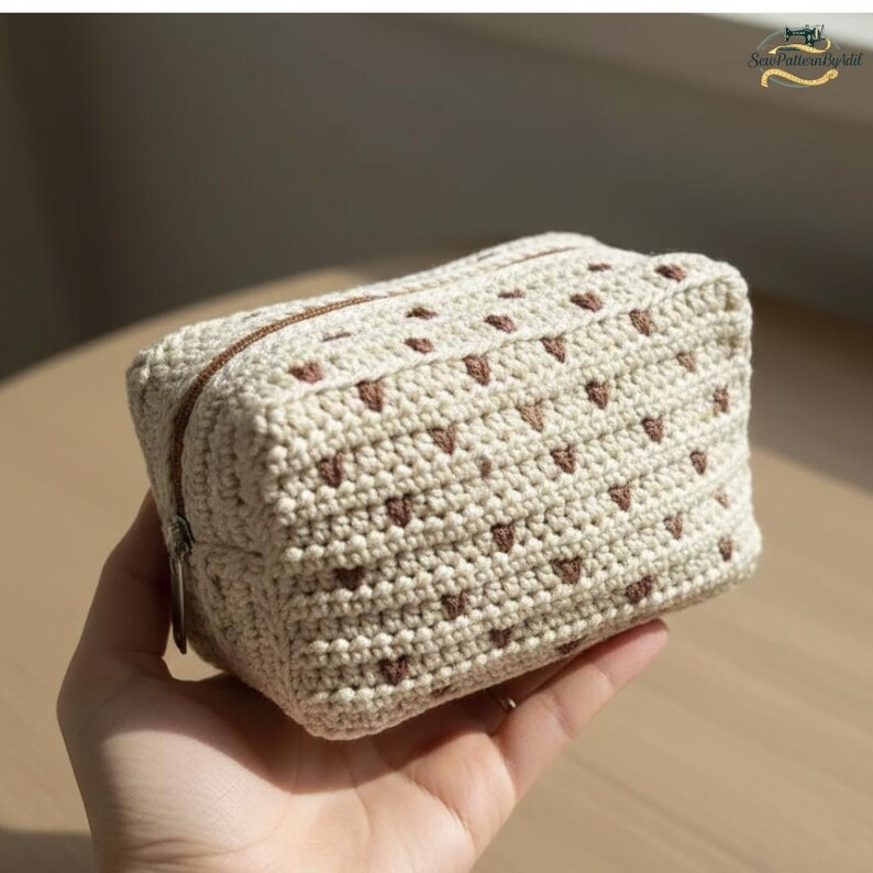 May include: A small, cream-colored crochet pouch with a brown zipper and heart-shaped accents. The handmade bag is held in a hand and is likely used for storing small items. The crochet stitches are visible, and the overall design is simple and charming.