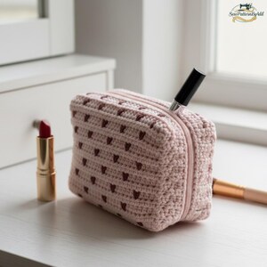 May include: A light pink crochet cosmetic bag with a heart pattern and a zipper closure. A lipstick and a makeup brush are next to the bag. A black lipstick tube is sticking out of the zipper.