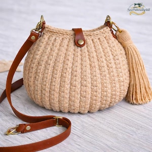 May include: A beige crochet crossbody bag with a brown leather strap and tassel. The bag has a rounded shape with vertical ribbed texture. The strap is adjustable and attaches with gold-colored hardware. The bag is closed with a leather flap and a gold button.
