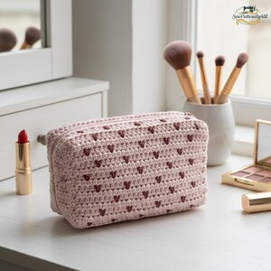 May include: A light pink, rectangular crochet cosmetic bag with a zipper closure. The bag is adorned with small, dark pink heart-shaped patterns. The bag is sitting on a white surface, next to makeup brushes, lipstick, and a mirror.