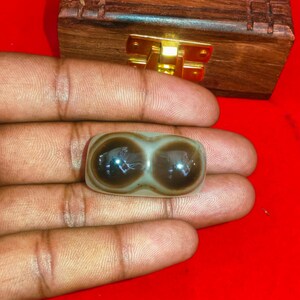 Puede incluir: Una piedra pulida, de forma ovalada, con dos marcas marrones oscuras en forma de ojo. La piedra es de color gris claro con remolinos marrones y se sostiene en una mano. Una caja de madera con un cierre dorado está en el fondo.