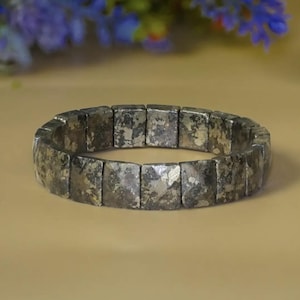 May include: A stretch bracelet composed of rectangular, metallic-toned pyrite beads. Each bead features a textured, reflective surface with mottled grey and gold-coloured inclusions, arranged in a continuous circular band without a visible clasp.