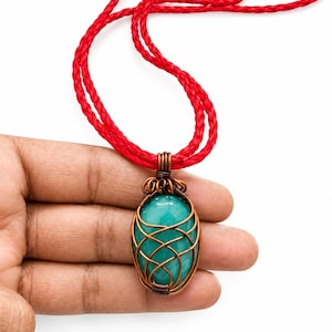 Pode incluir: Um colar com pingente apresentando uma pedra oval e lisa de cor verde-azulado, envolta em uma gaiola decorativa de fio de cobre. O pingente está suspenso por um cordão trançado vermelho brilhante que forma um laço no topo, criando contraste.