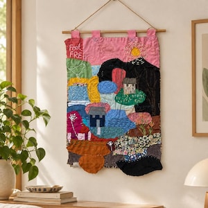 Puede incluir: Un tapiz rectangular de tela con un colorido mosaico de escenas bordadas, que incluye una casa, árboles y un sol amarillo. El texto "Feel FREE" está cosido en la esquina superior izquierda. Cuelga de una varilla de madera con pestañas rosas.