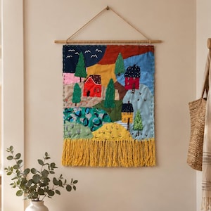 Puede incluir: Un tapiz rectangular de tela que presenta un paisaje bordado colorido con casas, árboles y colinas. La pieza textil está suspendida de una varilla de madera con un cordel y terminada con un fleco largo y grueso de color amarillo en el borde inferior.