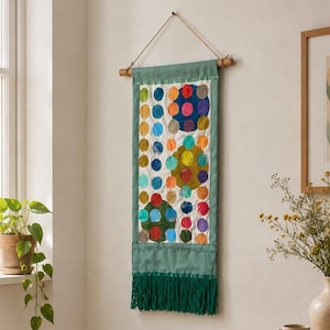 Puede incluir: Un tapiz rectangular con un panel de tela blanca decorado con un patrón de círculos multicolores en tonos azul, verde, amarillo, naranja y rojo. El panel está enmarcado por un borde verde salvia y rematado con flecos de color verde oscuro.