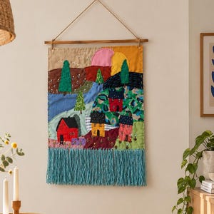 Puede incluir: Un tapiz rectangular de tela con un paisaje que incluye casas bordadas en rojo, amarillo y rosa. El diseño presenta árboles verdes, un río azul y un sol con semicírculos rosas y amarillos. Tiene un largo fleco de color verde azulado en la parte inferior.