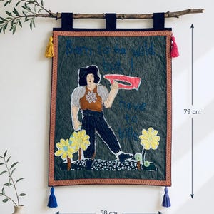 May include: A rectangular quilted wall hanging featuring an embroidered figure, yellow flowers, and the text "Born to be wild but I have to bills". The textile art measures 79 cm in height and 58 cm in width, accented with colourful tassels.