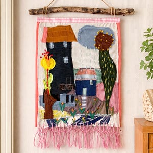 Könnte beinhalten: Textil-Wandbehang mit einer skurrilen Landschaftsszene. Das Kunstwerk zeigt Häuser, Bäume und eine Blume, die alle aus verschiedenen Stoffen gefertigt sind. Das Stück hat einen rosa Rand und eine Fransen am unteren Rand und ist an einem Holzstab aufgehängt.