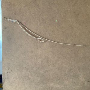 May include: A close-up of the rear of a framed artwork. The artwork is secured to a brown, textured backing board with staples and twine. The twine is looped for hanging.
