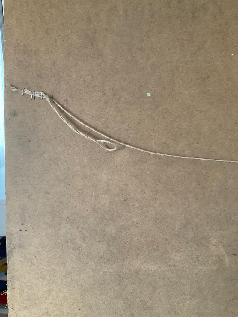 May include: A close-up of the rear of a framed artwork. The artwork is secured to a brown, textured backing board with staples and twine. The twine is looped for hanging.