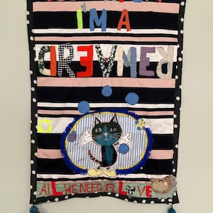 Könnte beinhalten: Ein Wandbehang aus Stoff mit dem Text "YOU MAY SAY I'M A DREAMER" in bunten Buchstaben. Darunter eine Cartoon-Katze und die Worte "ALL WE NEED IS LOVE". Der Banner hat schwarz-weiße Punkte und blaue Quasten.