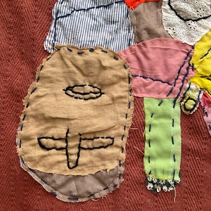 Könnte beinhalten: Textilkunstwerk mit einem Patchwork-Design. Das Kunstwerk zeigt verschiedene Stoffreste in Farben wie Braun, Rot, Blau, Rosa und Gelb. Die Teile sind mit kontrastierendem Faden zusammengenäht, wodurch ein strukturierter und geschichteter Effekt entsteht. Das Design beinhaltet eine gesichtsähnliche Form.