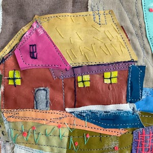 Op de afbeelding: Een textielkunstwerk met een huis met een geel dak, roze gevel en roodbruine muren. Het huis heeft gele ramen met blauwe kruissteekjes. Het omliggende landschap bevat oranje, groene en blauwe stoffen lappen met roze geborduurde bloemen en blauwe decoratieve stiksels.