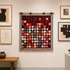 Op de afbeelding: Een wandkleed met een raster van cirkelvormige stoffen lapjes in rood, zwart, blauw en wit, opgehangen aan een houten roede met een grijze franjerand. Het werk hangt aan een witte muur, omringd door diverse ingelijste kunstwerken.