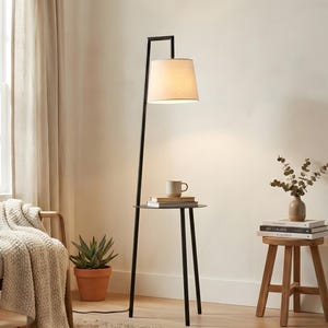 Puede incluir: Una lámpara de pie de metal negro con una mesa auxiliar redonda. La lámpara tiene una pantalla beige y un soporte vertical delgado. Una pequeña mesa sostiene una taza y libros. Una planta en maceta y un taburete de madera también están en la habitación.