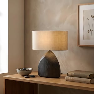 Peut inclure: Une lampe de table moderne avec un socle texturé gris foncé et un abat-jour rectangulaire beige. La lampe est posée sur une table en bois, à côté d'un petit bol avec des pierres et deux livres. Le design de la lampe est contemporain et minimaliste.