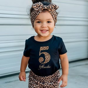 Può includere: Un bambino indossa un completo composto da maglietta e pantaloncini neri con un motivo leopardato. Il numero "5" e il nome "Annika" sono stampati sulla maglietta con stampa leopardata. Una fascia abbinata con stampa leopardata completa l'outfit.