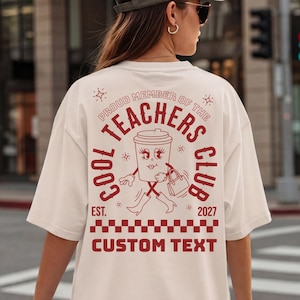 Könnte beinhalten: Ein cremefarbenes Kurzarm-T-Shirt mit einem roten Aufdruck auf der Rückseite. Das Motiv zeigt eine Comic-Kaffeetasse mit Gesicht, Stiefeln und Handtasche, umgeben von dem Text "PROUD MEMBER OF THE COOL TEACHERS CLUB EST. 2027" und "CUSTOM TEXT".