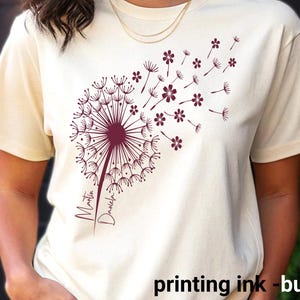 Könnte beinhalten: Cremefarbenes T-Shirt mit einem bordeauxfarbenen Pusteblumen-Design. Die Samen der Pusteblume fliegen davon, mit den Namen "Martin" und "Daniela" in Schreibschrift. Der Text "printing ink - burgundy" steht unten.