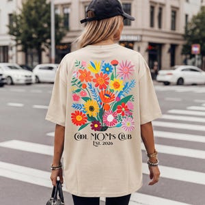 Könnte beinhalten: Beiges T-Shirt mit einem bunten Blumenstrauß-Design auf dem Rücken. Die Grafik zeigt verschiedene Blumen in Rot-, Gelb-, Blau- und Rosatönen. Der Text "COOL MOM'S CLUB EST. 2026" ist darunter gedruckt.