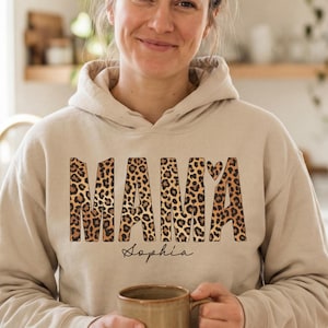 Può includere: Felpa con cappuccio beige con la scritta "MAMA" in stampa leopardata, un cuore e il nome "Sophia" in corsivo. La persona tiene in mano una tazza marrone.