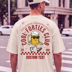 Könnte beinhalten: Ein cremefarbenes T-Shirt mit einer Grafik auf der Rückseite, die einen gezeichneten Bierkrug mit Sonnenbrille und Turnschuhen zeigt. Der Text lautet "EXCLUSIVE MEMBERS ONLY", "COOL FORTIES CLUB", "EST. 1986" und "CUSTOM TEXT" über einem rot-weißen Karomuster.