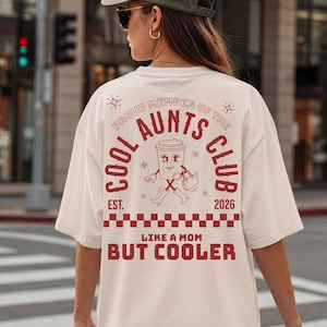 Könnte beinhalten: Ein cremefarbenes T-Shirt mit einem roten Aufdruck auf der Rückseite. Der Text lautet PROUD MEMBER OF THE COOL AUNTS CLUB EST. 2026 und LIKE A MOM BUT COOLER. Die Grafik zeigt einen Kaffeebecher mit Gesicht, Armen, Beinen und einer Handtasche.