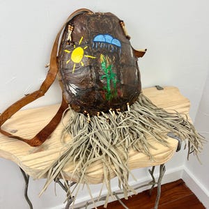 May include: A brown leather bag made from a turtle shell with a fringe trim. The bag has a leather strap and is decorated with painted designs of a sun, clouds, and a cactus.