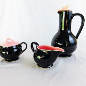 May include: A black ceramic tea set with pink accents. The set includes a teapot, creamer, and sugar bowl.