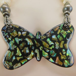 Puede incluir: Un collar de plata con un gran colgante de mariposa hecho de concha de abulón iridiscente. La mariposa está hecha de muchas piezas pequeñas de concha de abulón que están engastadas en una resina negra.
