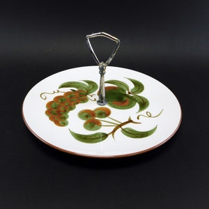 May include: A white ceramic serving tray with a brown rim and a silver handle. The tray features a painted design of green leaves, brown grapes, and green and orange apples.
