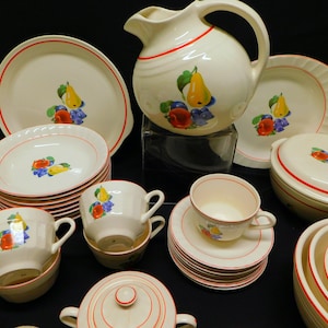 Può includere: Un set vintage di piatti in ceramica bianca con bordo rosso e un motivo a frutta. Il set comprende una brocca, ciotole, piatti e una zuccheriera.