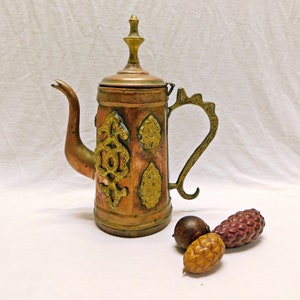 May include: A copper and brass teapot with ornate gold designs. The teapot has a spout and a handle. The teapot is sitting on a white surface with three small brown and gold pinecones.