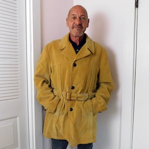 May include: Mustard yellow corduroy coat with a belted waist and button closure. The coat has a classic design with a notched collar and pockets. The coat is a mid-length style.