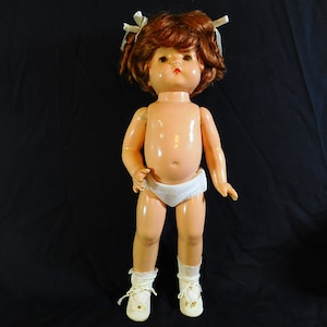 Puede incluir: Una muñeca de plástico vintage con cabello rojo, que lleva ropa interior blanca y zapatos blancos. La muñeca tiene una cara pintada con ojos marrones y labios rojos.