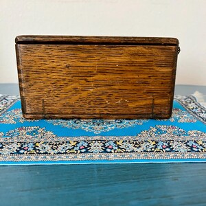 May include: A small, rectangular wooden box with a hinged lid. The box is made of a light brown wood with visible grain and some minor imperfections. It sits on a small, ornate blue rug.