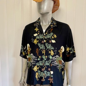 Puede incluir: Camisa negra con botones con un estampado vibrante de temática submarina. El diseño presenta sirenas, caballitos de mar, peces y corales en tonos verdes, marrones y dorados. La camisa tiene mangas cortas y un cuello clásico.