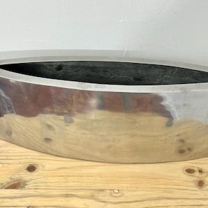 Op de afbeelding: Een gepolijste, metallic zilveren plantenbak met een bootachtige vorm. De plantenbak heeft een brede, ovale opening en staat op een houten oppervlak. De binnenkant is donker en contrasteert met de glanzende buitenkant.