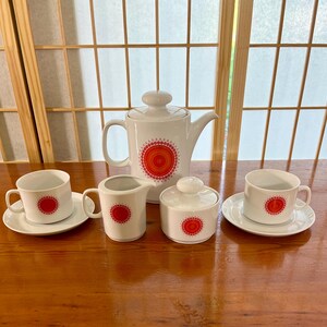 Puede incluir: Un juego de té blanco con un patrón geométrico rojo y naranja. El juego incluye una tetera, una jarra de crema, un azucarero y dos tazas de té y platillos.