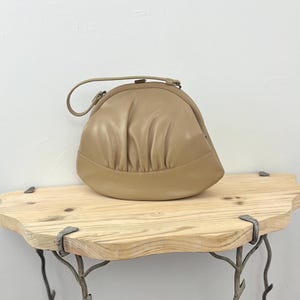 Puede incluir: Un bolso de mano de cuero marrón con forma curva y redondeada y un cierre de metal. El bolso está sobre una estantería de madera con un marco de metal decorativo.