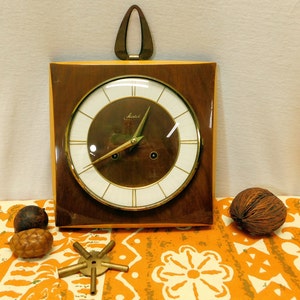 Può includere: Un orologio da parete quadrato in legno con un quadrante e lancette dorate. L'orologio ha una cornice in legno marrone e un coperchio in vetro trasparente. Il quadrante dell'orologio ha uno sfondo bianco con indicatori delle ore romani neri e una lancetta dei secondi dorata. L'orologio è realizzato dal marchio "Jauch".