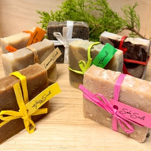 May include: Assortment of handmade soap bars in various colors, each wrapped with a ribbon and a tag. The soaps are in shades of brown, beige, and green. Some tags have text. The soaps are arranged on a wooden surface with greenery in the background.