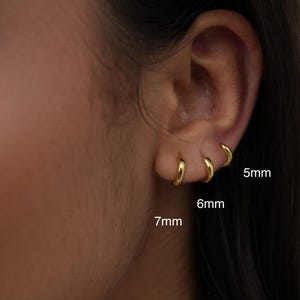 Pode incluir: Close-up de três brincos de argola dourados, tamanhos 5 mm, 6 mm e 7 mm, usados numa orelha. Os brincos são pequenos, circulares e brilhantes. O fundo é uma pele desfocada.